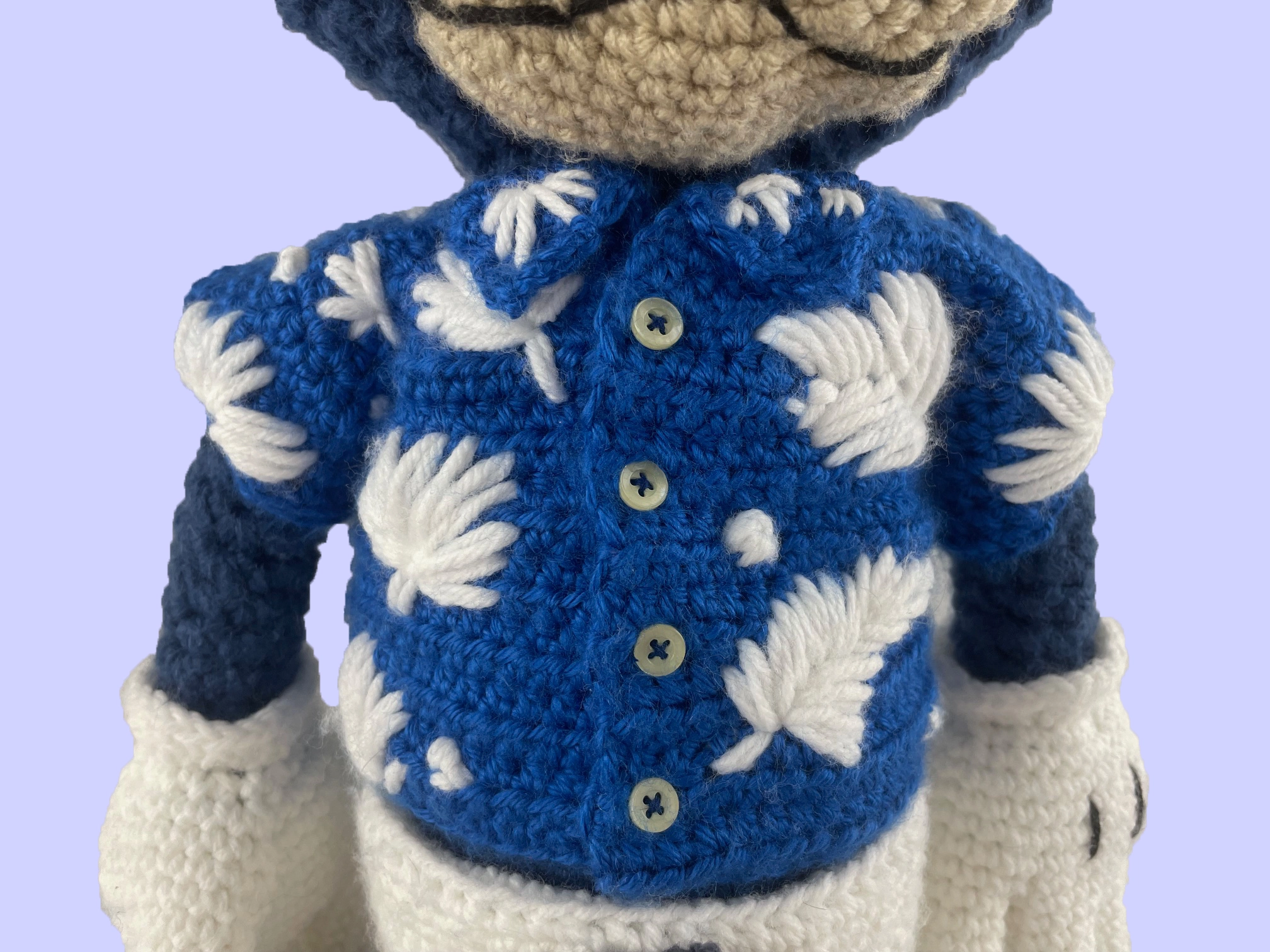Close-up of crochet Toontown blue cat plush wearing a handmade blue shirt with white floral pattern. Highlights stitching and outfit detail.